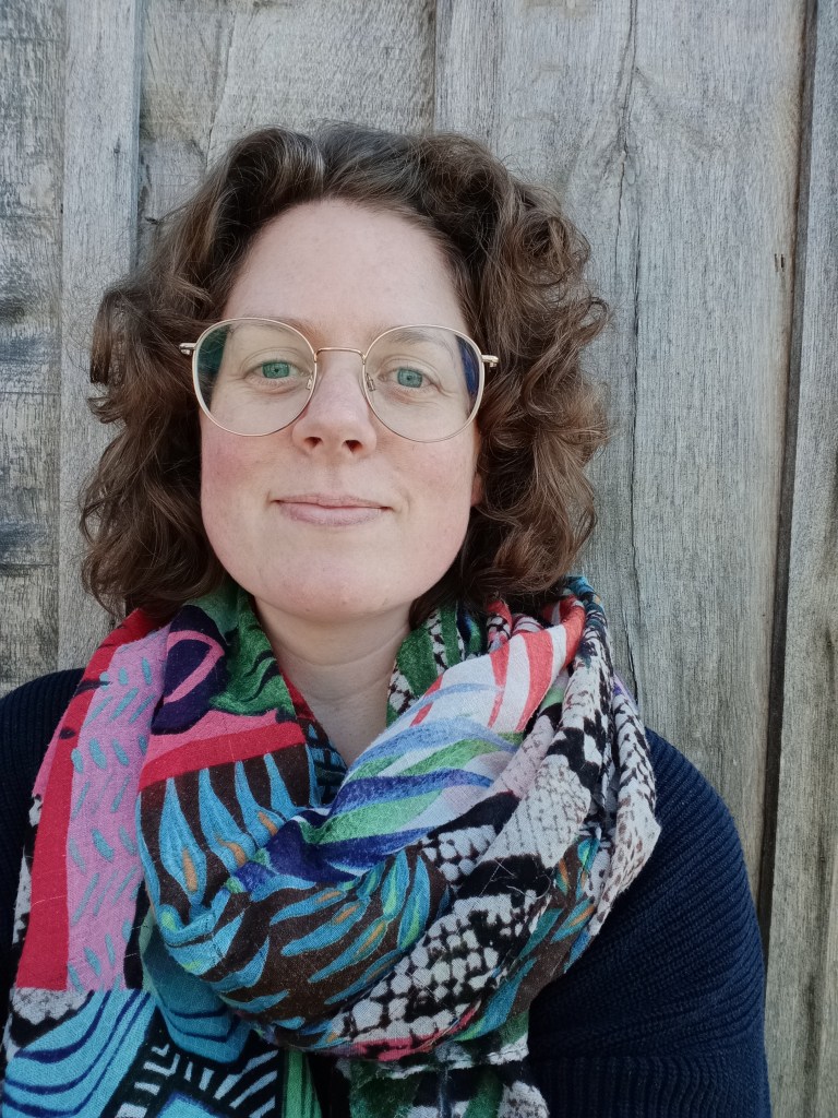Zelfportret. Een foto van een vrouw met bril, halflang krullend haar, blauwe trui en een gekleurde sjaal.