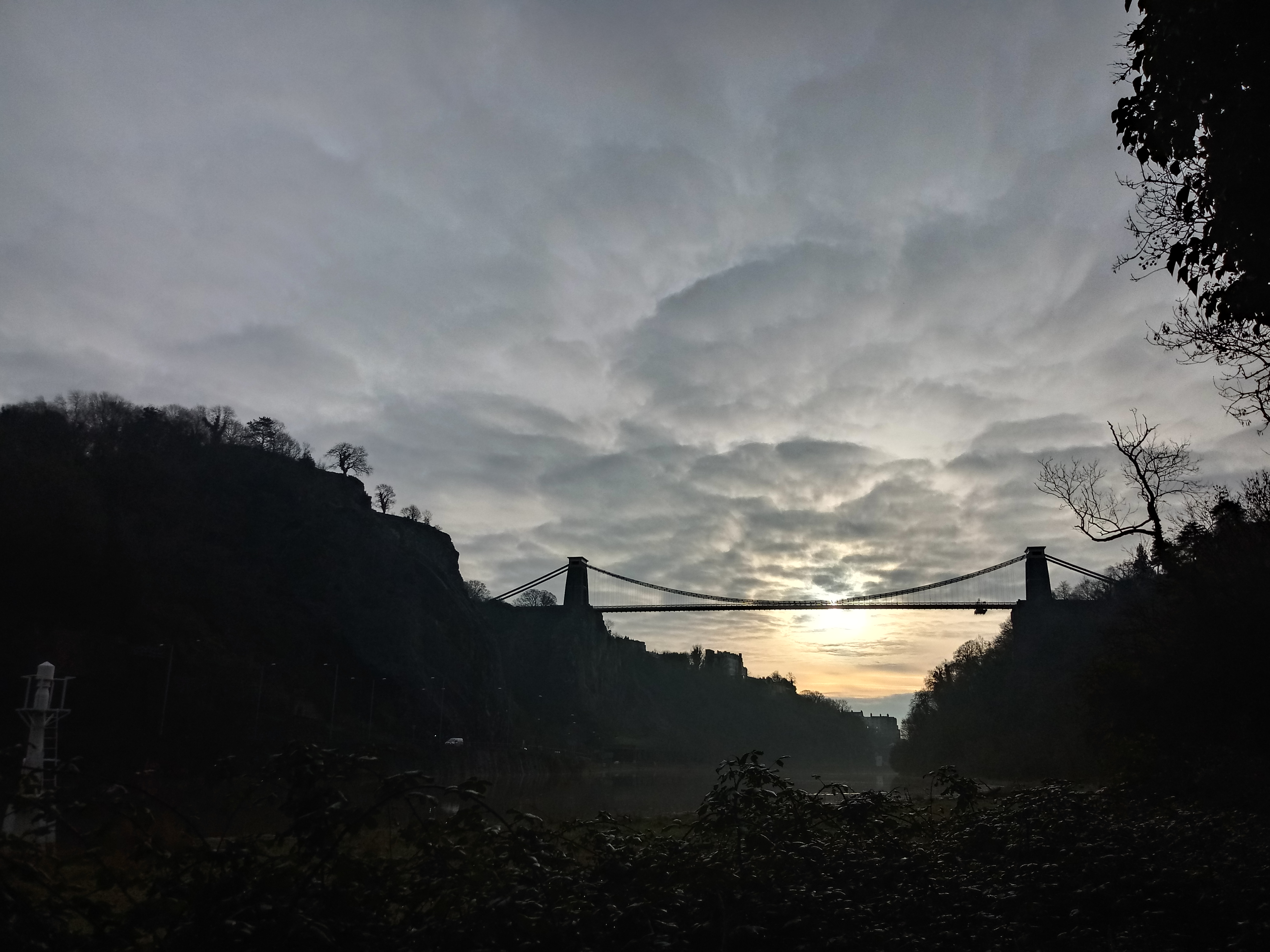Een donkere foto (kikvorsperspectief) van de Clifton Suspension Bridge waarbij in de verte de zonsondergang is te zien.