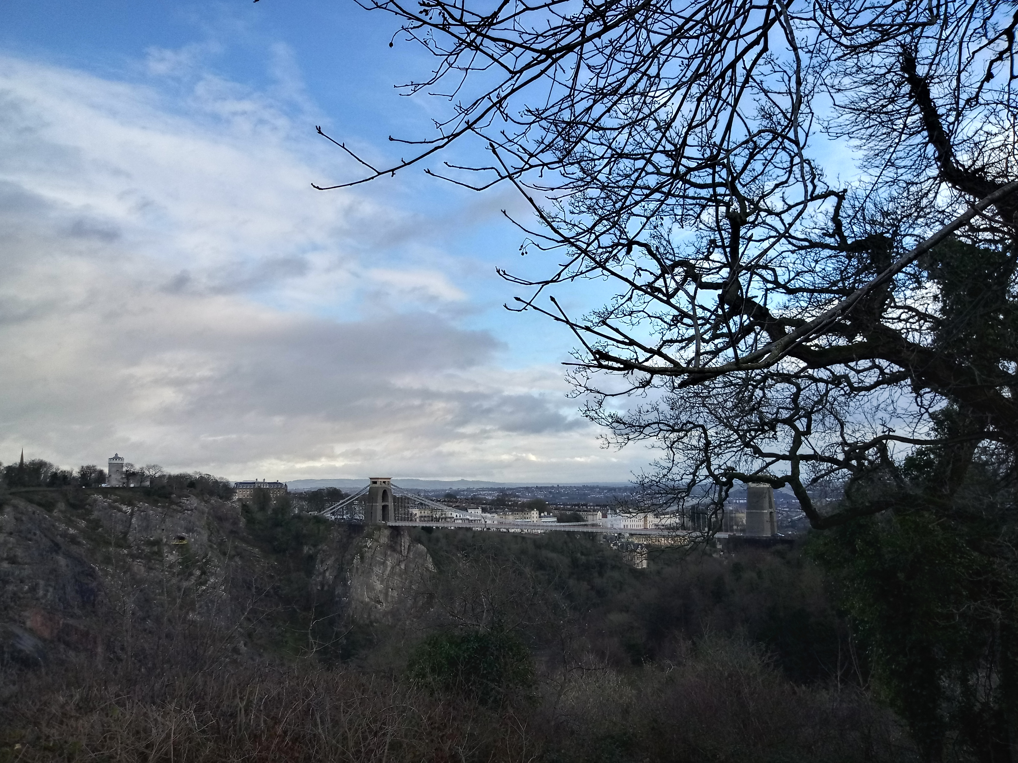 Tussen de takken van de bomen door is in de verte de Clifton Suspension Bridge zichtbaar. De lucht lijkt gesplitst in twee delen. Links de witte sluierbewolking, rechts blauwe lucht.