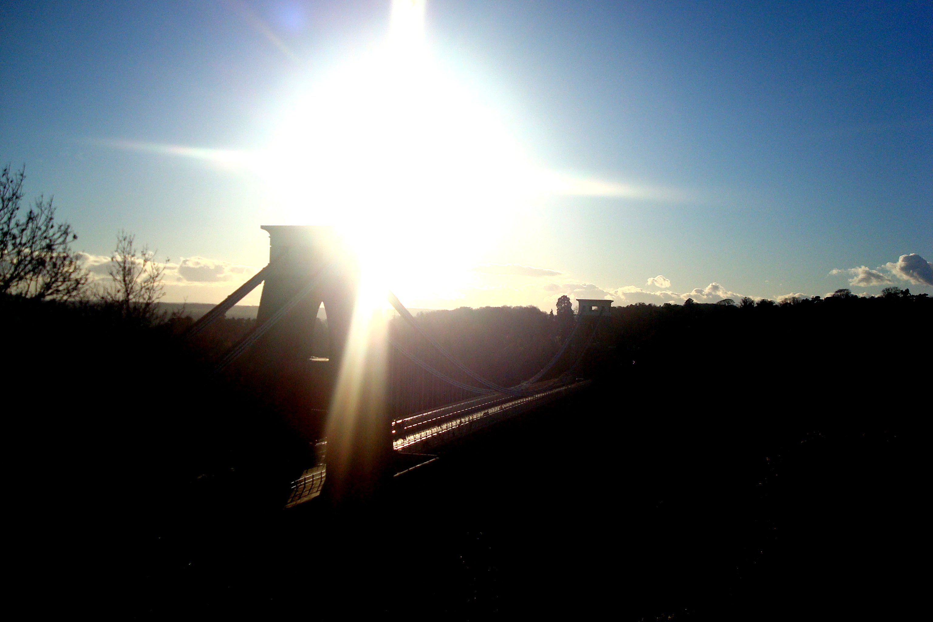 Een foto waarbij de zon hoog staat en valt over de Clifton Suspension Bridge. Door de felle zon is de vooral de omlijning van de brug zichtbaar.