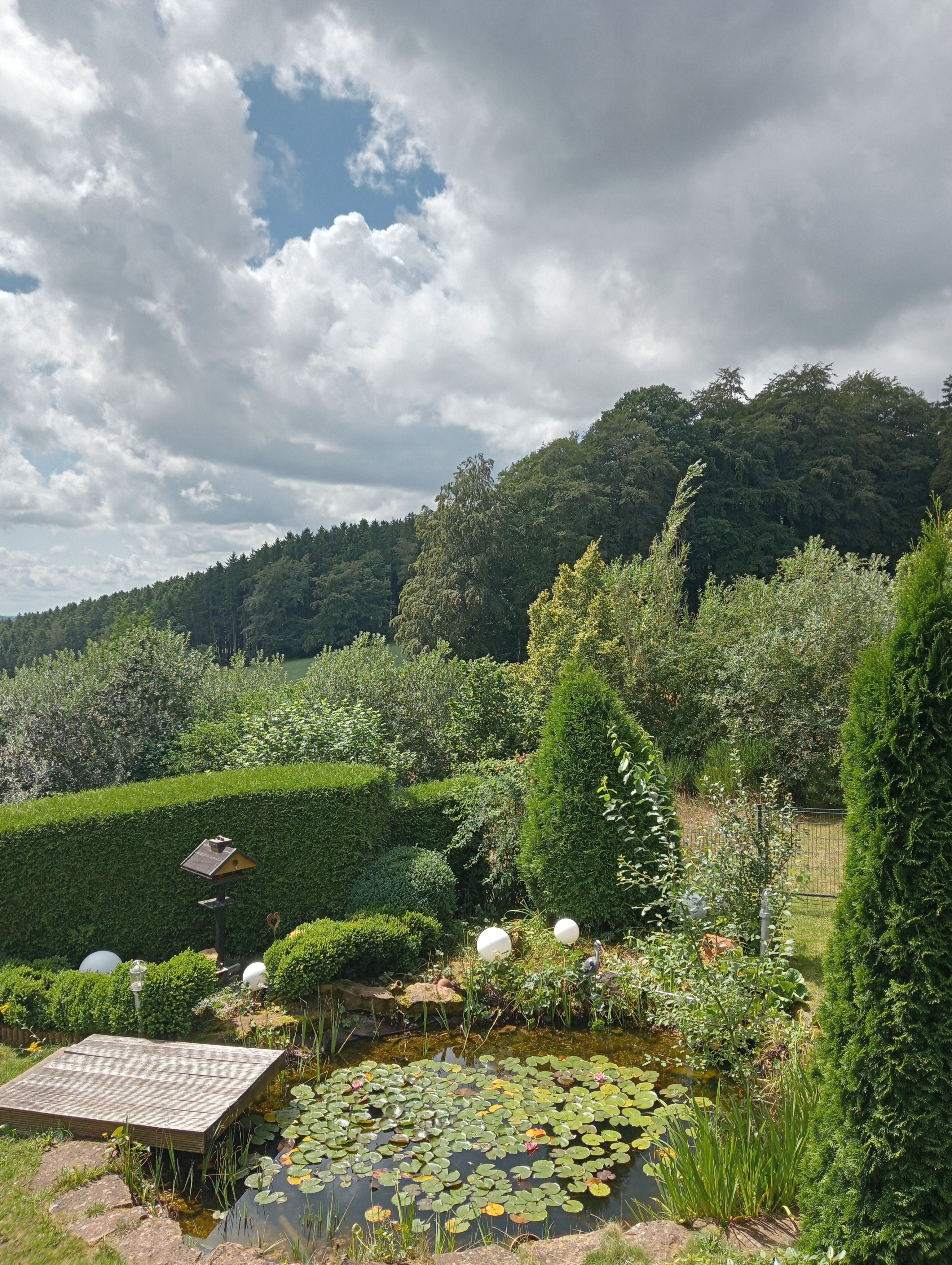 Een foto vanaf het balkon genomen met uitzicht op veel groen. Een vijver, vogelhuisje, een neppe reiger, groene heuvels en blauwe lucht met wolken.