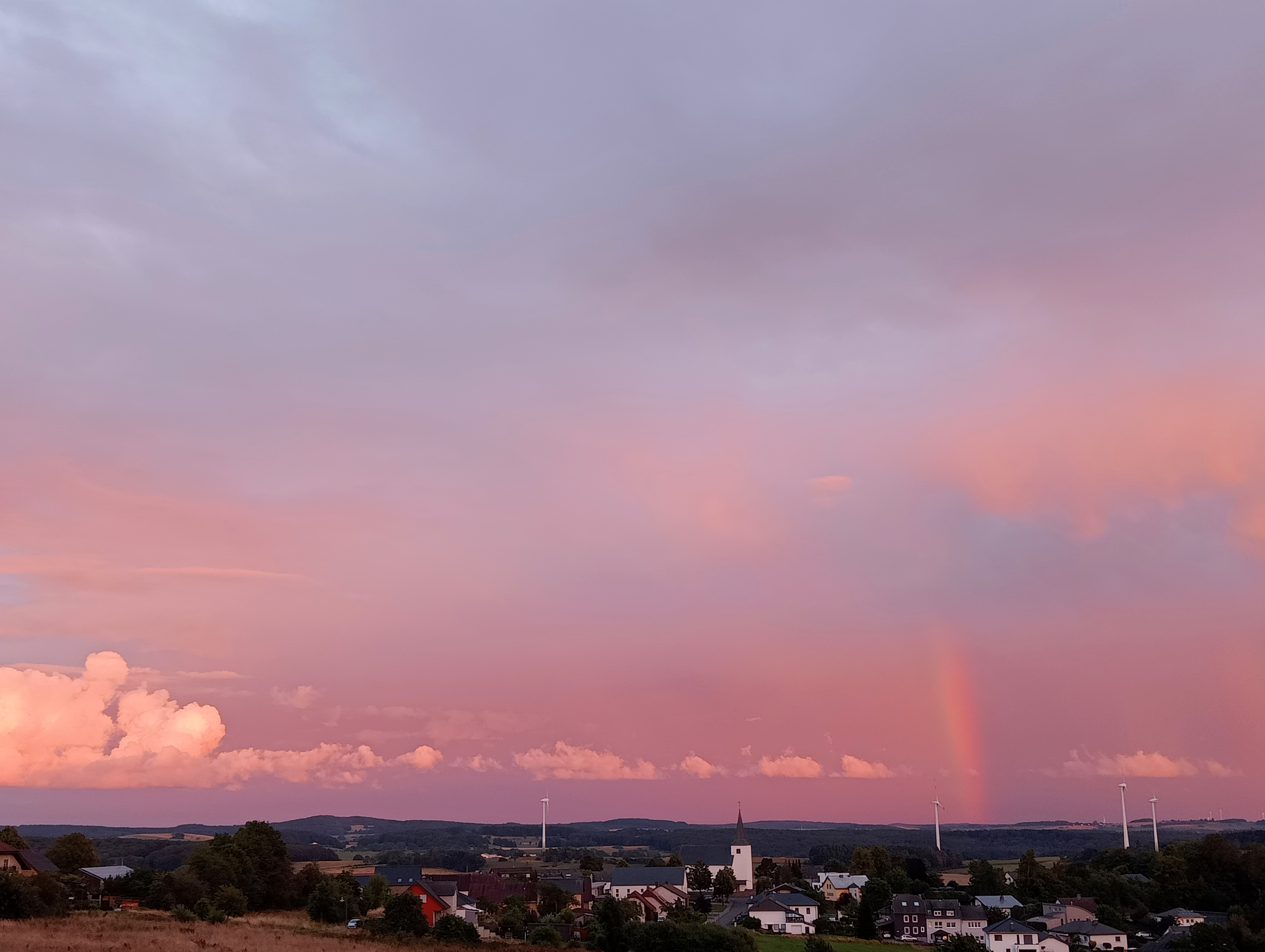 Verzicht. Een klein dorpje in de verte met een paars, oranje lucht daarboven en een regenboog die verschijnt.