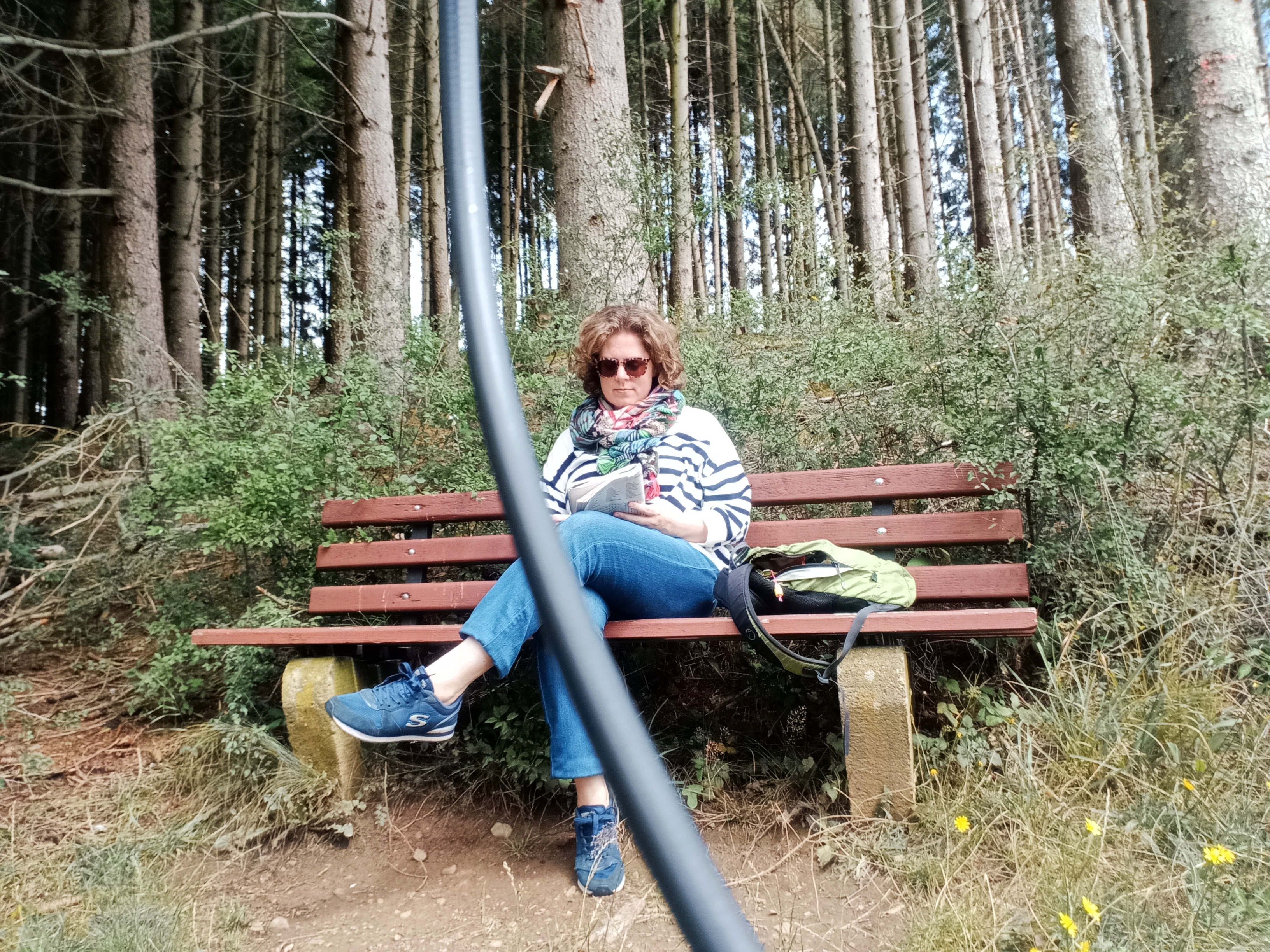 Een foto van mij waarbij ik op een bankje zit in natuurgebied Alfbachtal. Ik heb een zonnebril op en maak een woordzoeker. Achter mij staan hoge, groene bomen.