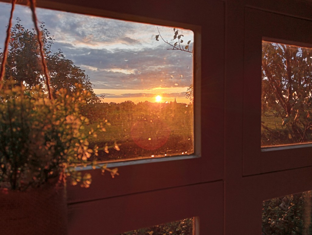 Foto door het raam van de pipowagen van binnen naar buiten met zicht op de zonsondergang die een prachtige gloed laat zien.